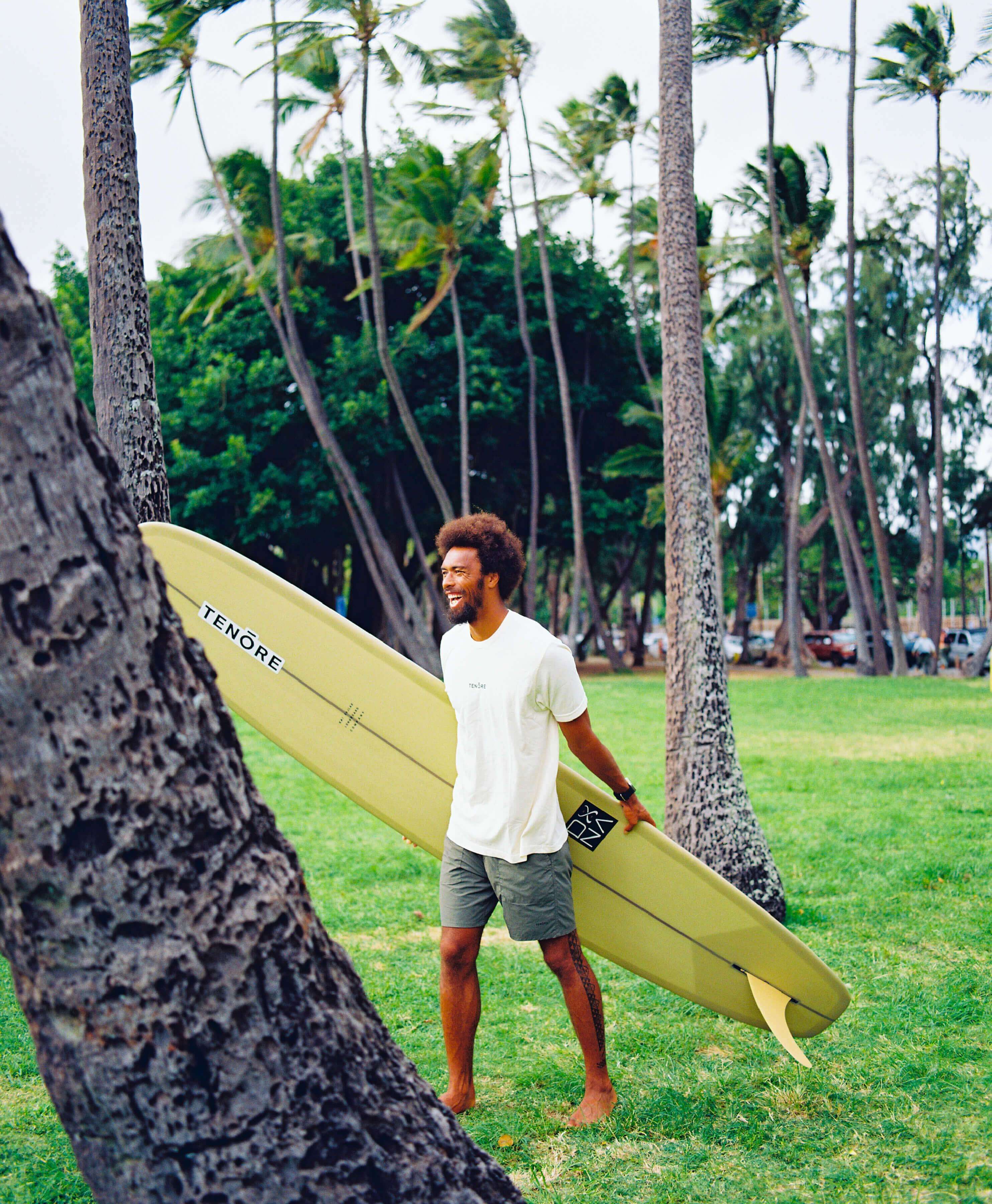 man holding yellow surfboard amongst palm trees and smiling