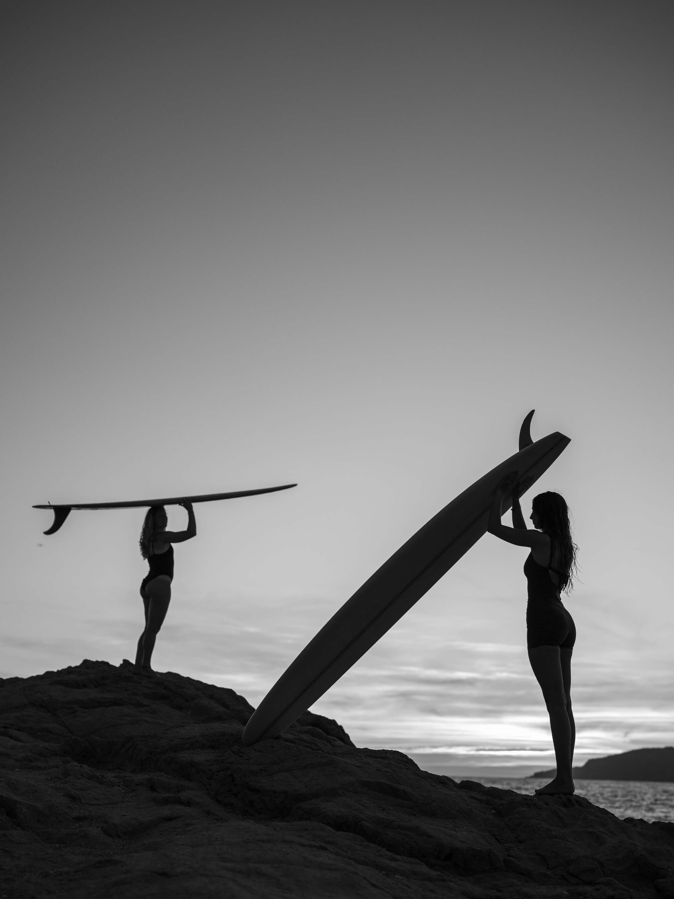 black and white silhouettes of two womans holding surfboards over their heads