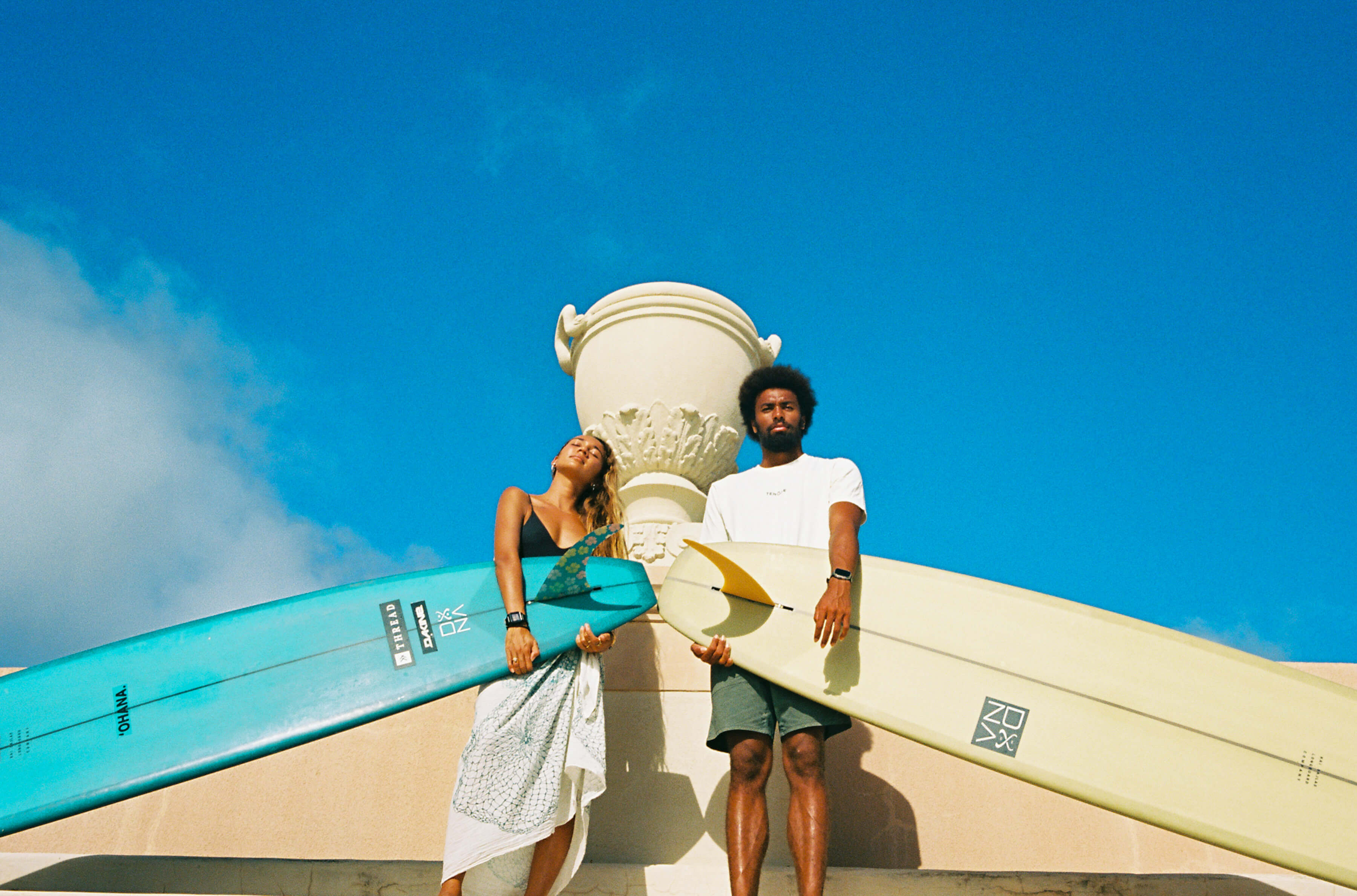 man and woman holding surfboards in front of white decorative urn