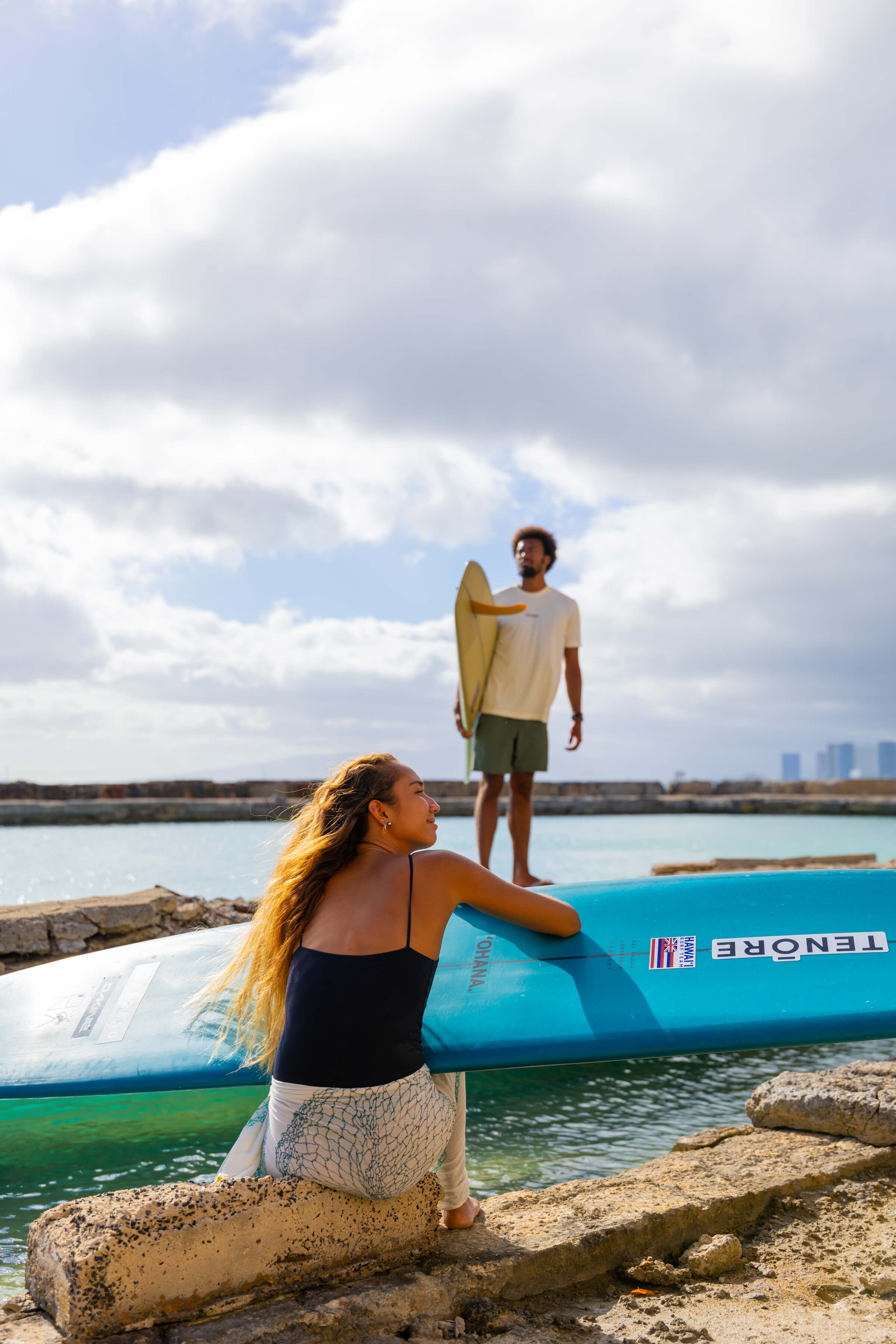 shot of woman with surfboard on lap looking into distance with man posing in background