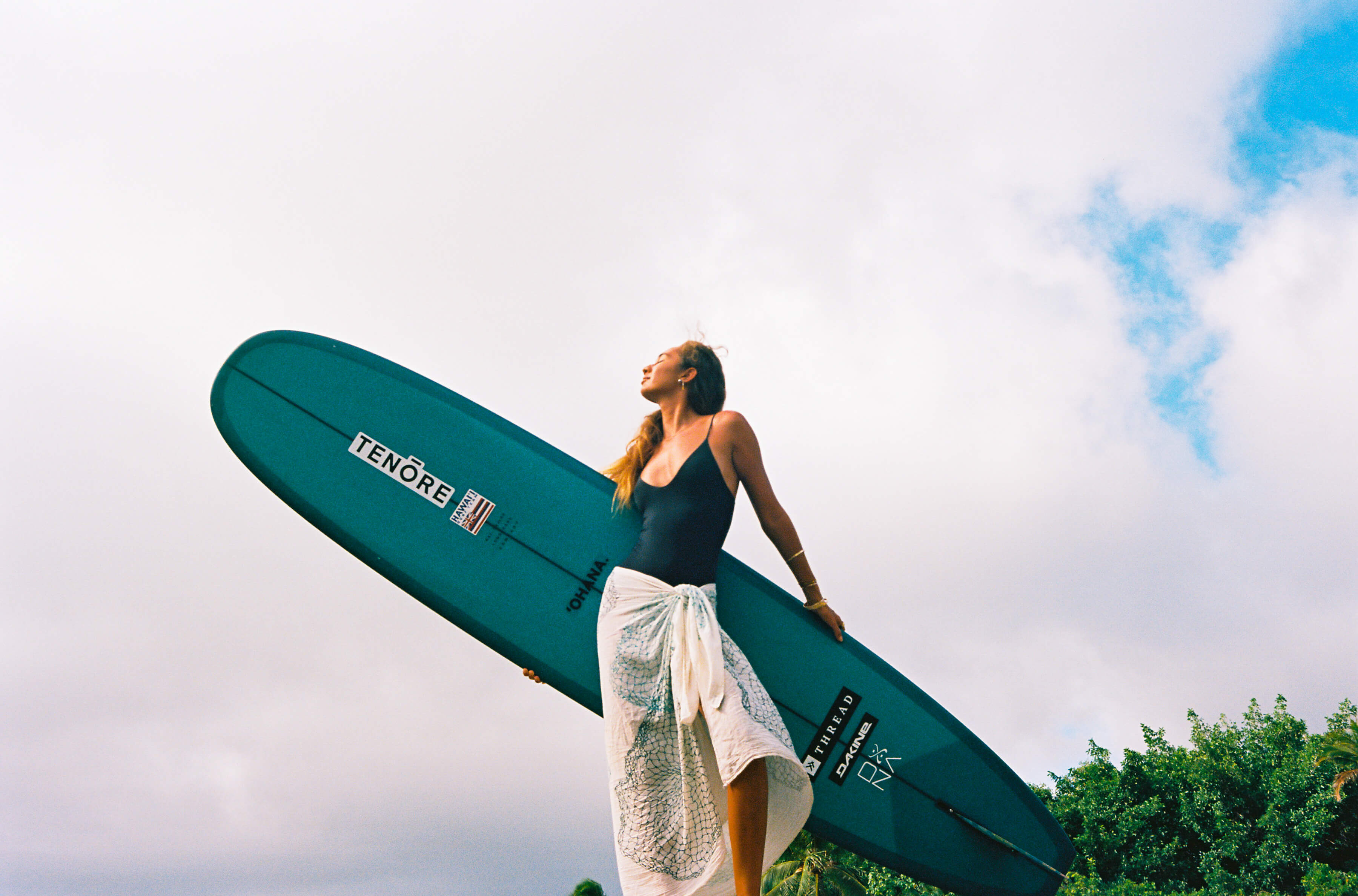 woman in flowy skirt holding surfboard behind her and looking to sky 