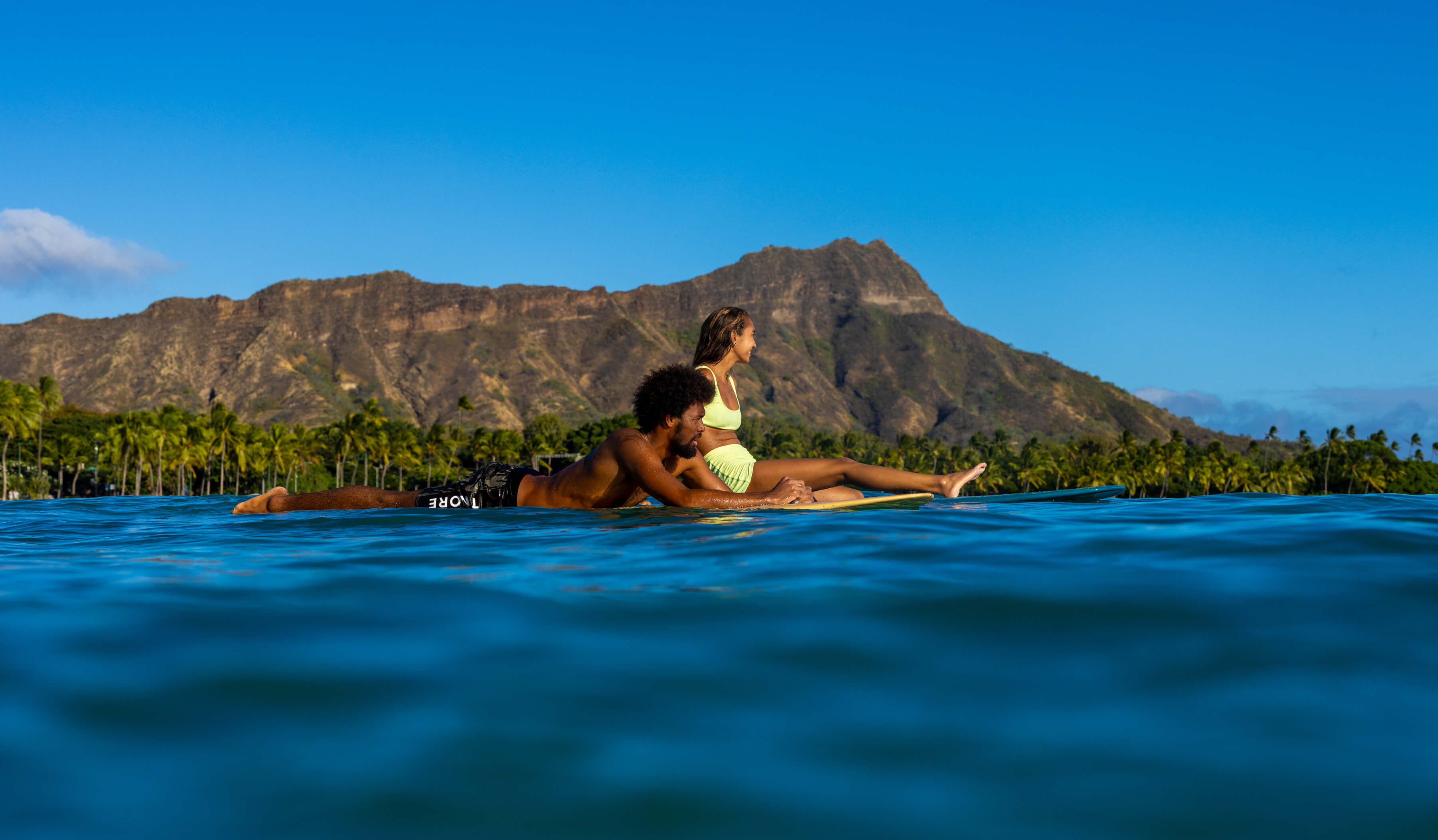 man and woman relaxing on surfboards in calm waters with hawaiian mountains in background