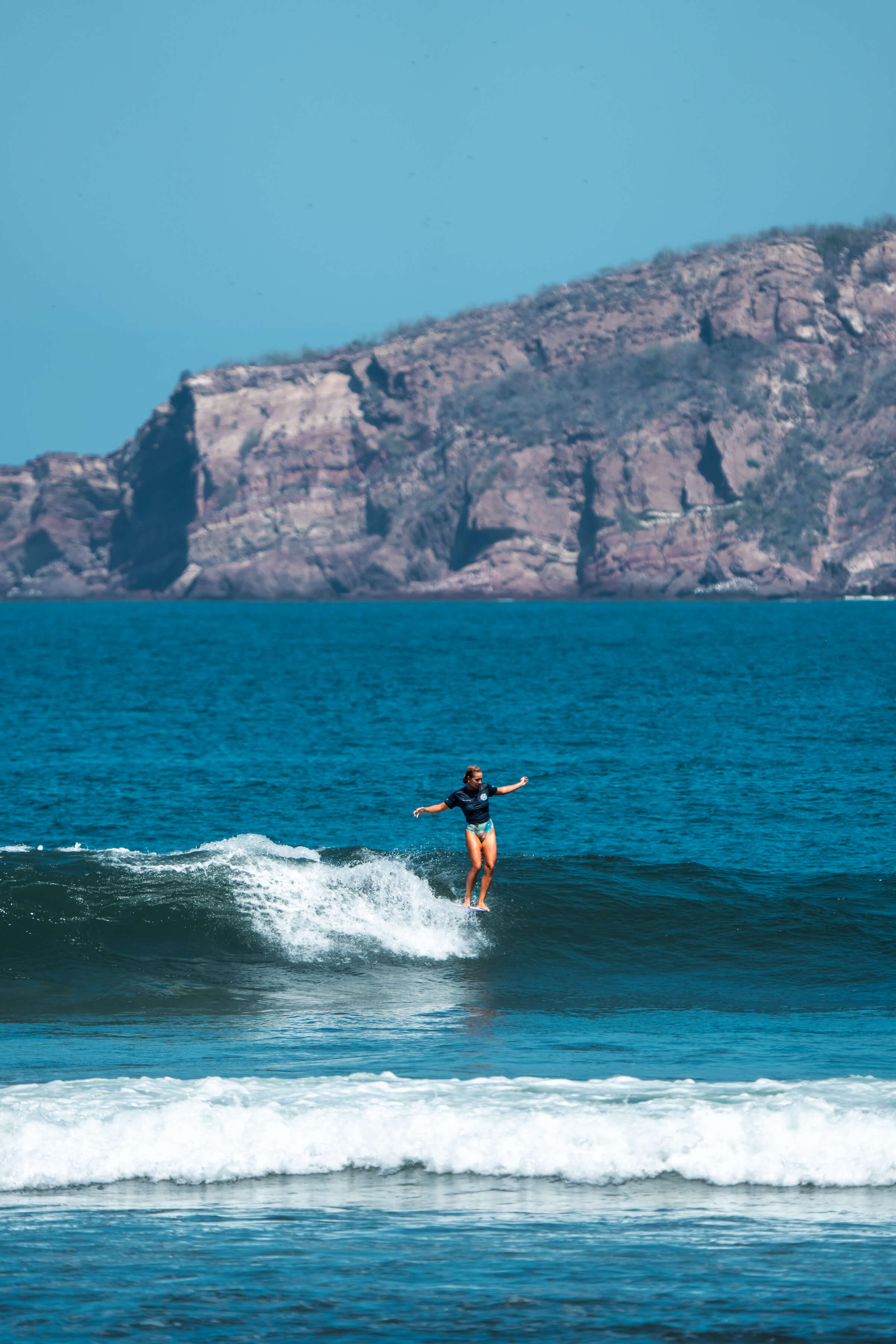 zoomed out view of woman riding wave with cliffs in background