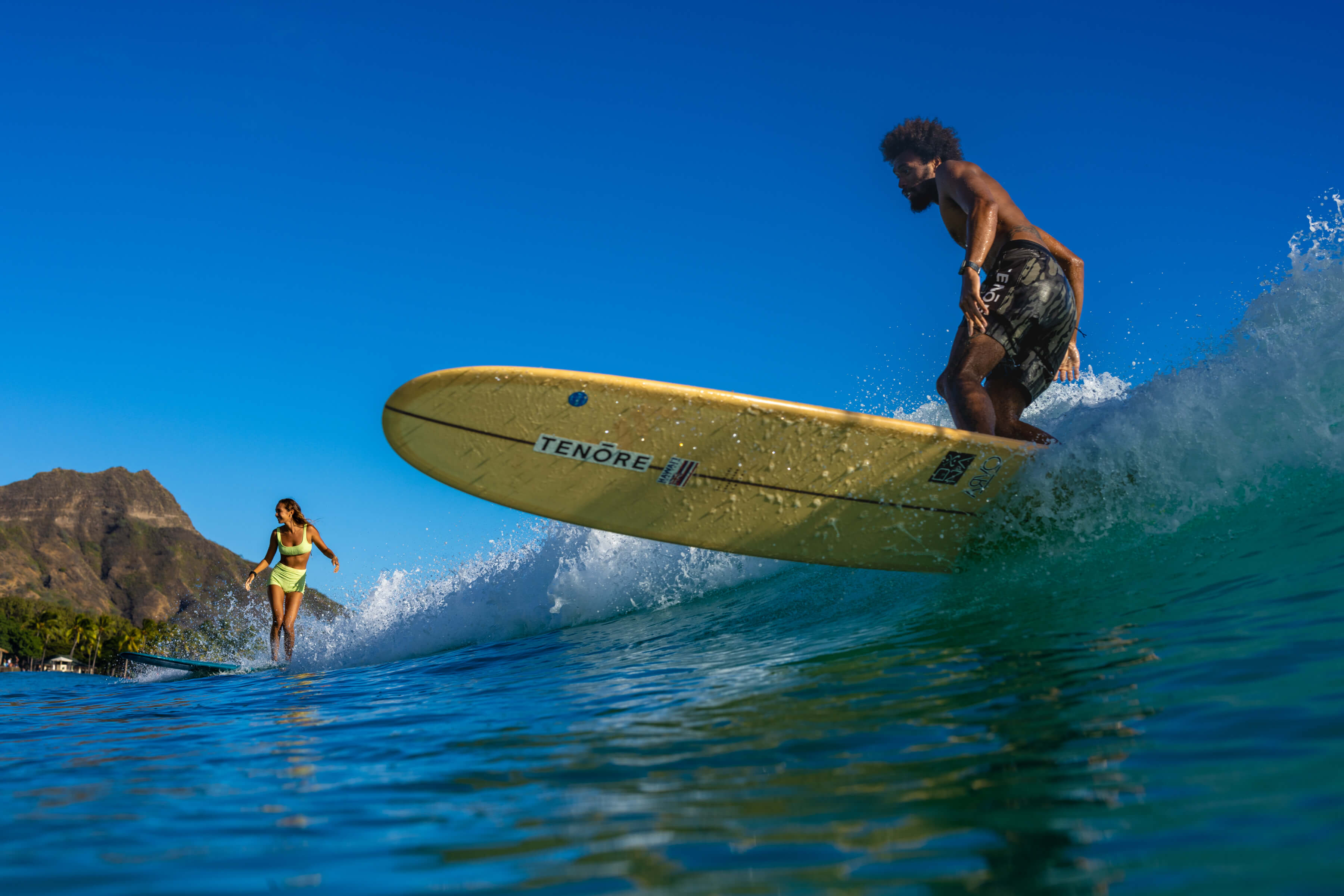 closeup of man on yellow surfboard riding wave with woman surfing behind him