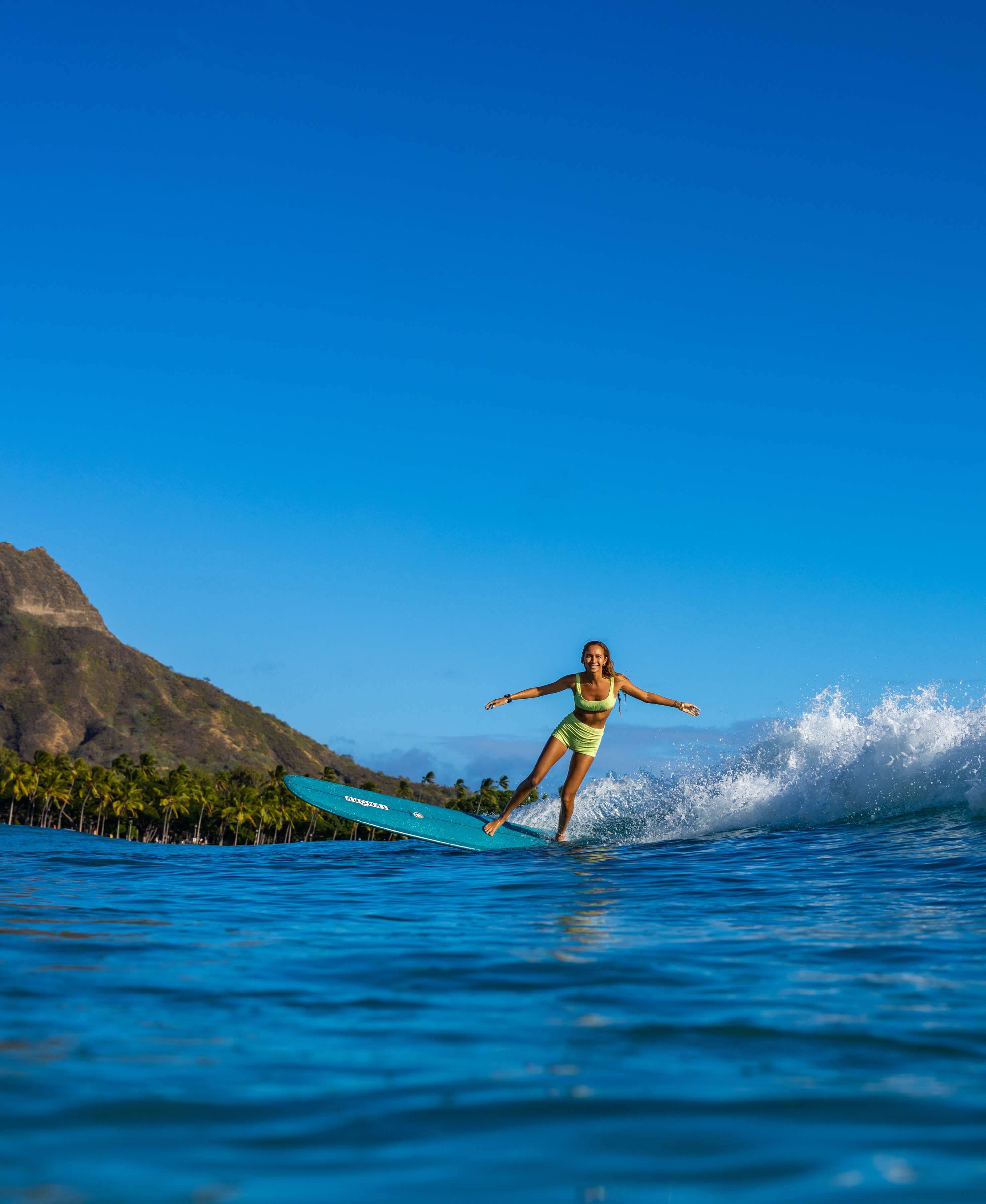 woman in yellow swim top and shorts smiling on a surfboard
