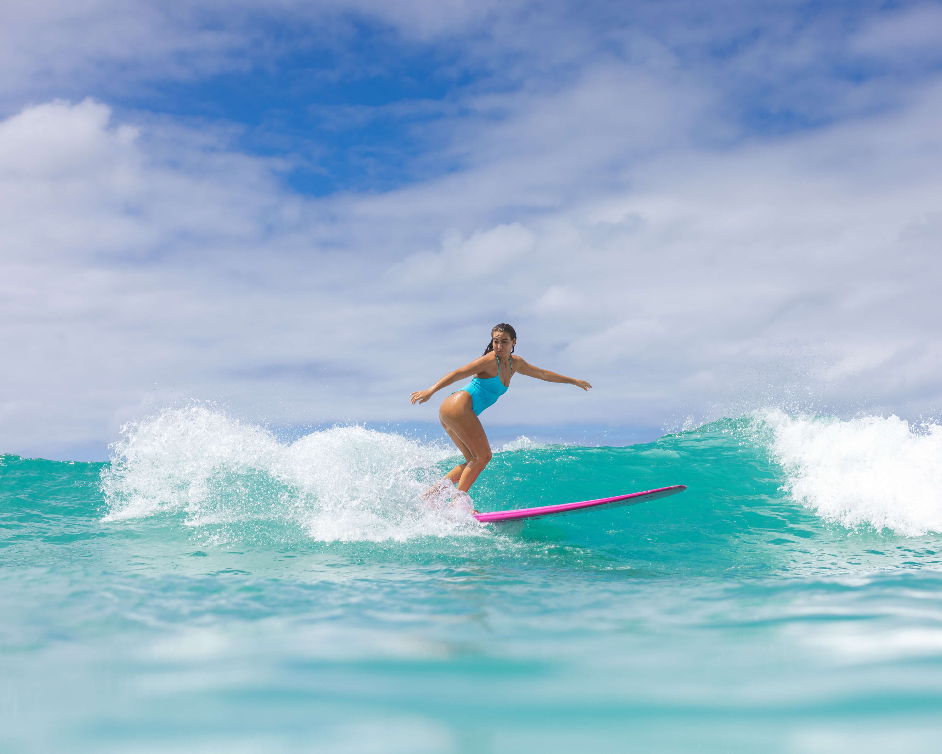 woman leaning on pink surfboard into wave with blue swimsuit
